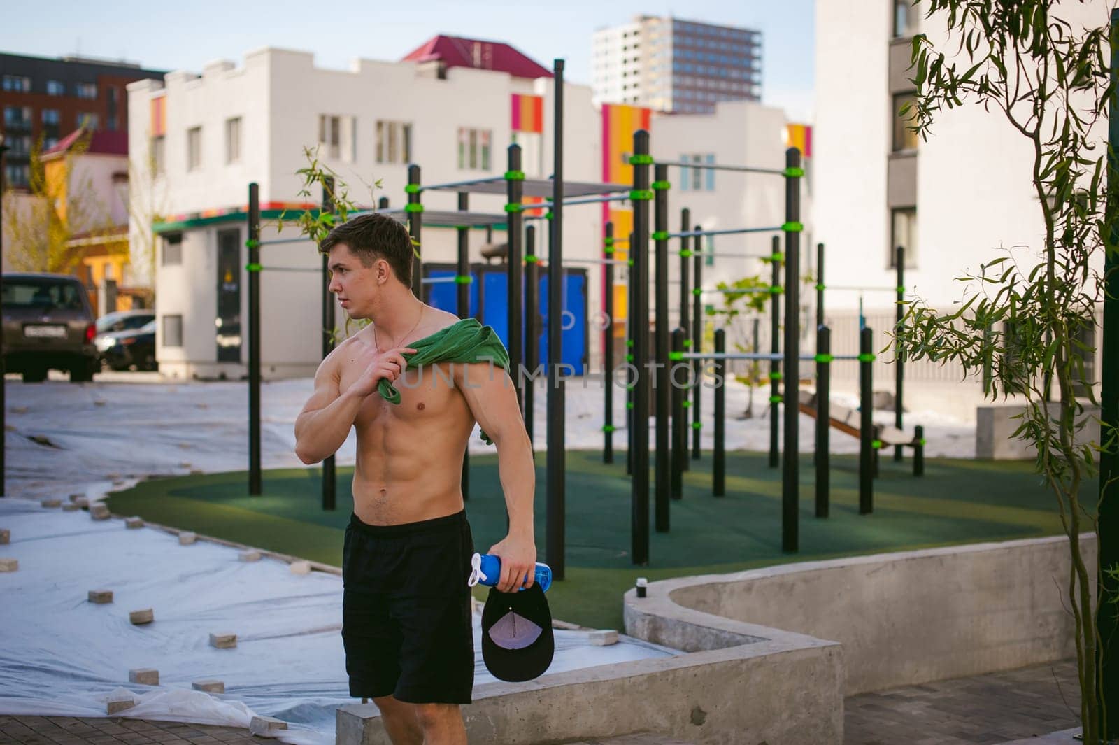 Handsome sexy male bodybuilder athlete man with naked torso, Poses showing a powerful muscular body standing near a green grid fence sports field in courtyard. Healthy lifestyle concept.