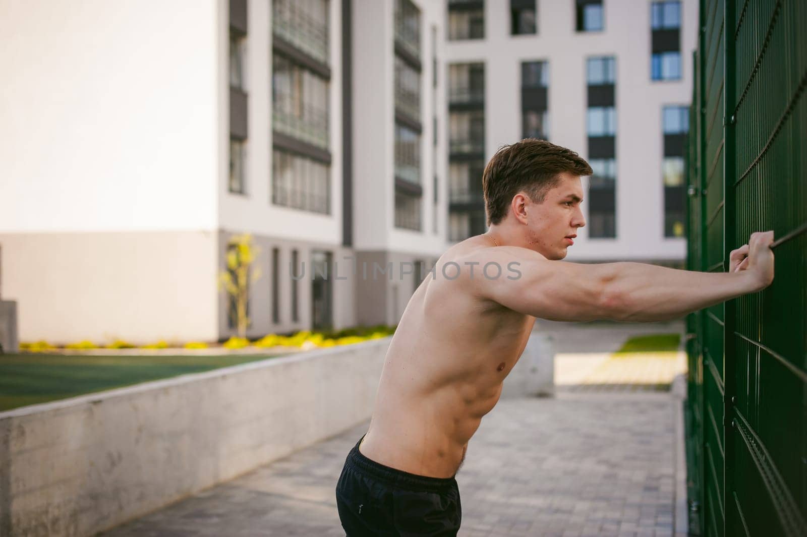 Handsome sexy male bodybuilder athlete man with naked torso, Poses showing a powerful muscular body standing near a green grid fence sports field in courtyard. Healthy lifestyle concept.