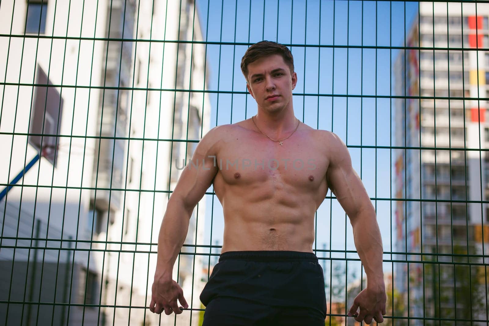 Handsome sexy male bodybuilder athlete man with naked torso, Poses showing a powerful muscular body standing near a green grid fence sports field in courtyard. Healthy lifestyle concept.