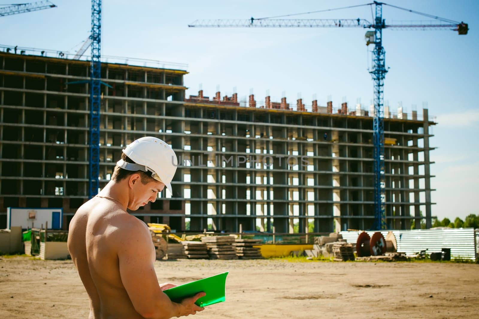 Young sexy male bodybuilder athlete with a bare torso. Manager engineer builder in white protective helmet stands on background of construction of building and construction cranes with documentation