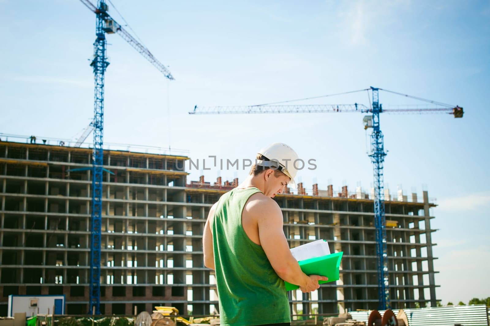 Young sexy men bodybuilder athlete. Manager engineer builder in white protective helmet stands on background of construction of building and construction cranes with documentation in hand