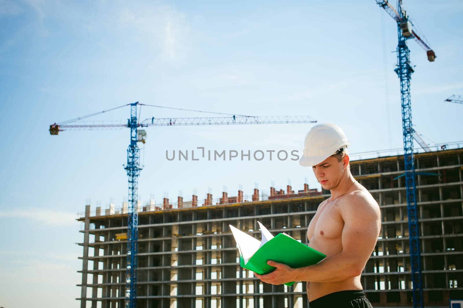 Young sexy male bodybuilder athlete with a bare torso. Manager engineer builder in white protective helmet stands on background of construction of building and construction cranes with documentation