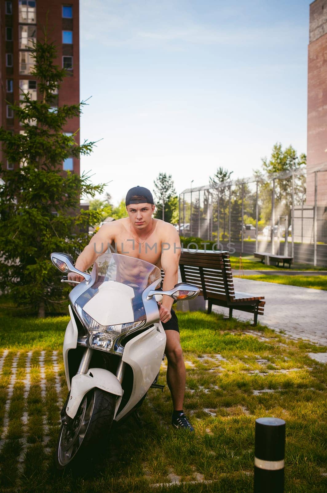 Handsome sexy male athlete man with naked torso in yard of house in parking lot sitting on a white sport motorcycle, an early sunny summer morning