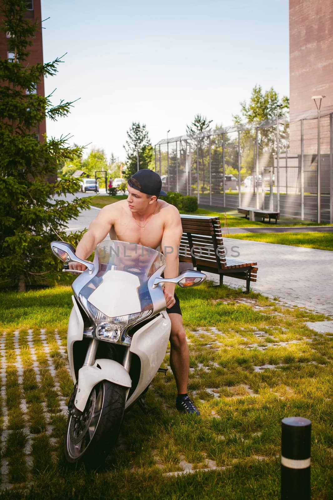 Handsome sexy male athlete man with naked torso in yard of house in parking lot sitting on a white sport motorcycle, an early sunny summer morning