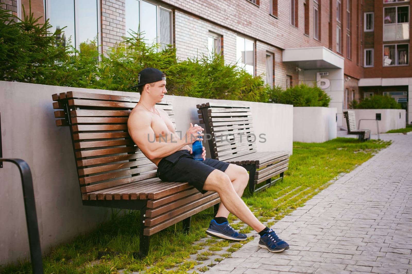 Handsome sexy male athlete man with naked torso resting on a bench in the courtyard of the house, after training drinks water from a bottle, an early sunny summer morning