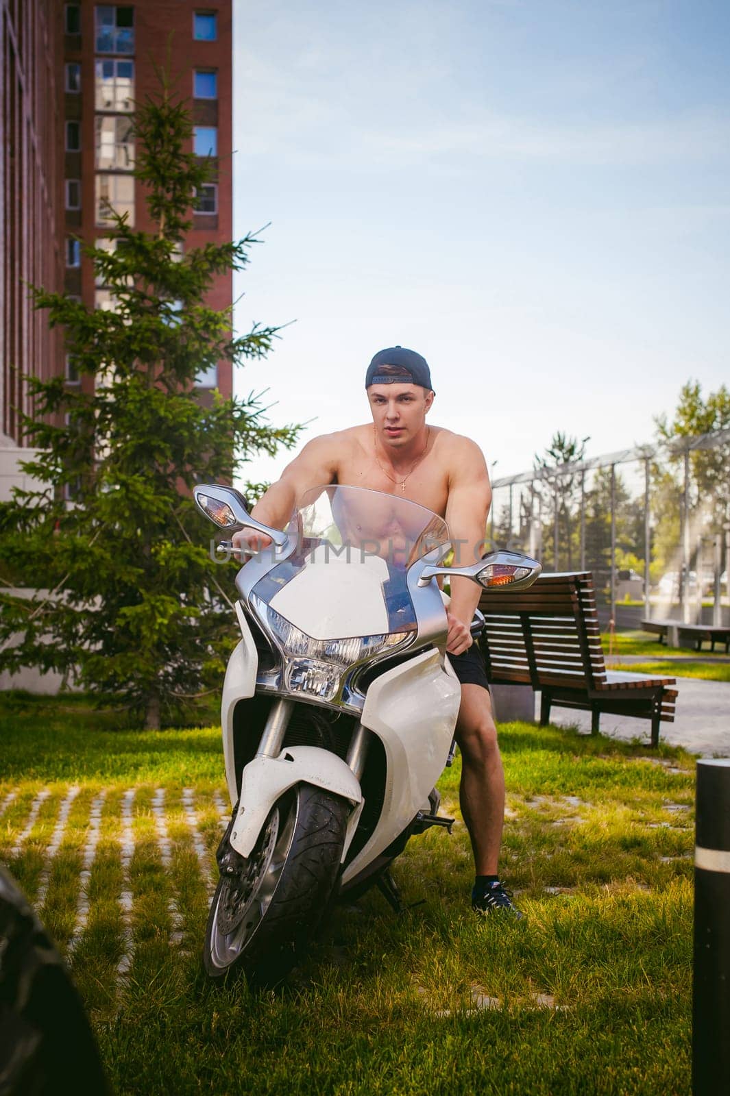 Handsome sexy male athlete man with naked torso in yard of house in parking lot sitting on a white sport motorcycle, an early sunny summer morning