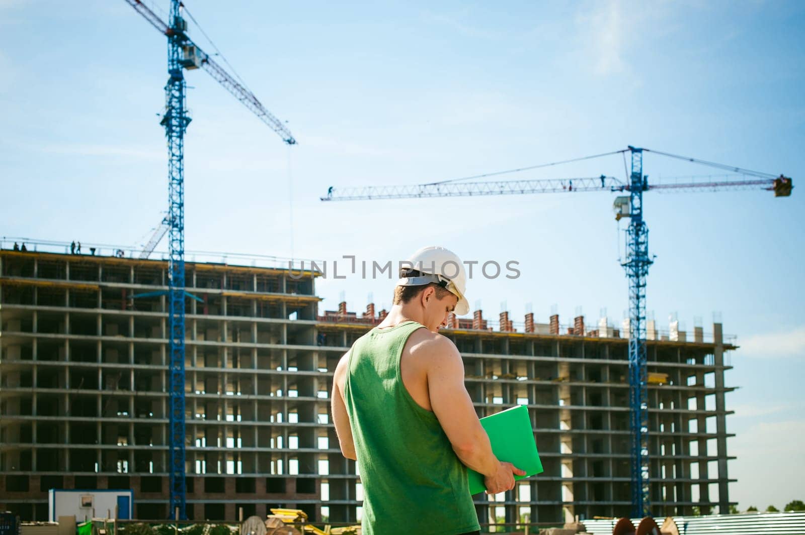 Young sexy men bodybuilder athlete. Manager engineer builder in white protective helmet stands on background of construction of building and construction cranes with documentation in hand