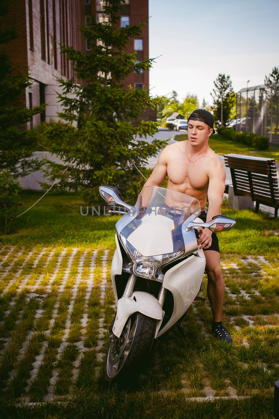Handsome sexy male athlete man with naked torso in yard of house in parking lot sitting on a white sport motorcycle, an early sunny summer morning