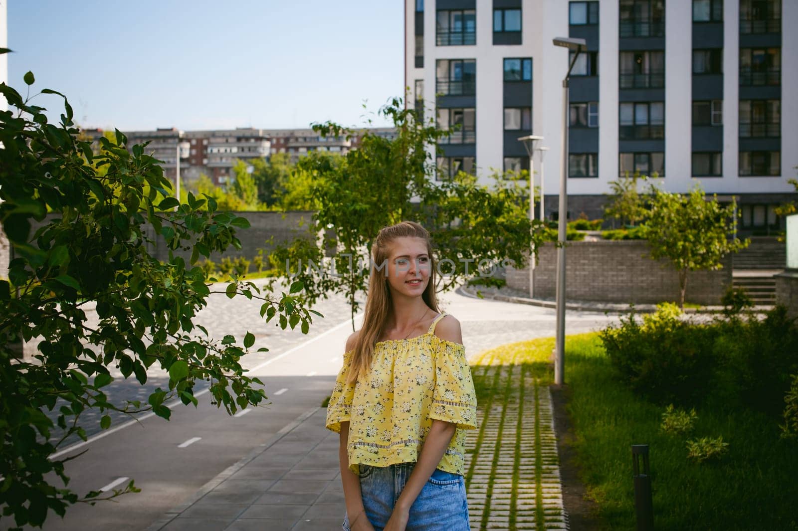 Young beautiful woman is strolling outdoors in the yard, against the backdrop of greenery and houses, warm summer sunny day