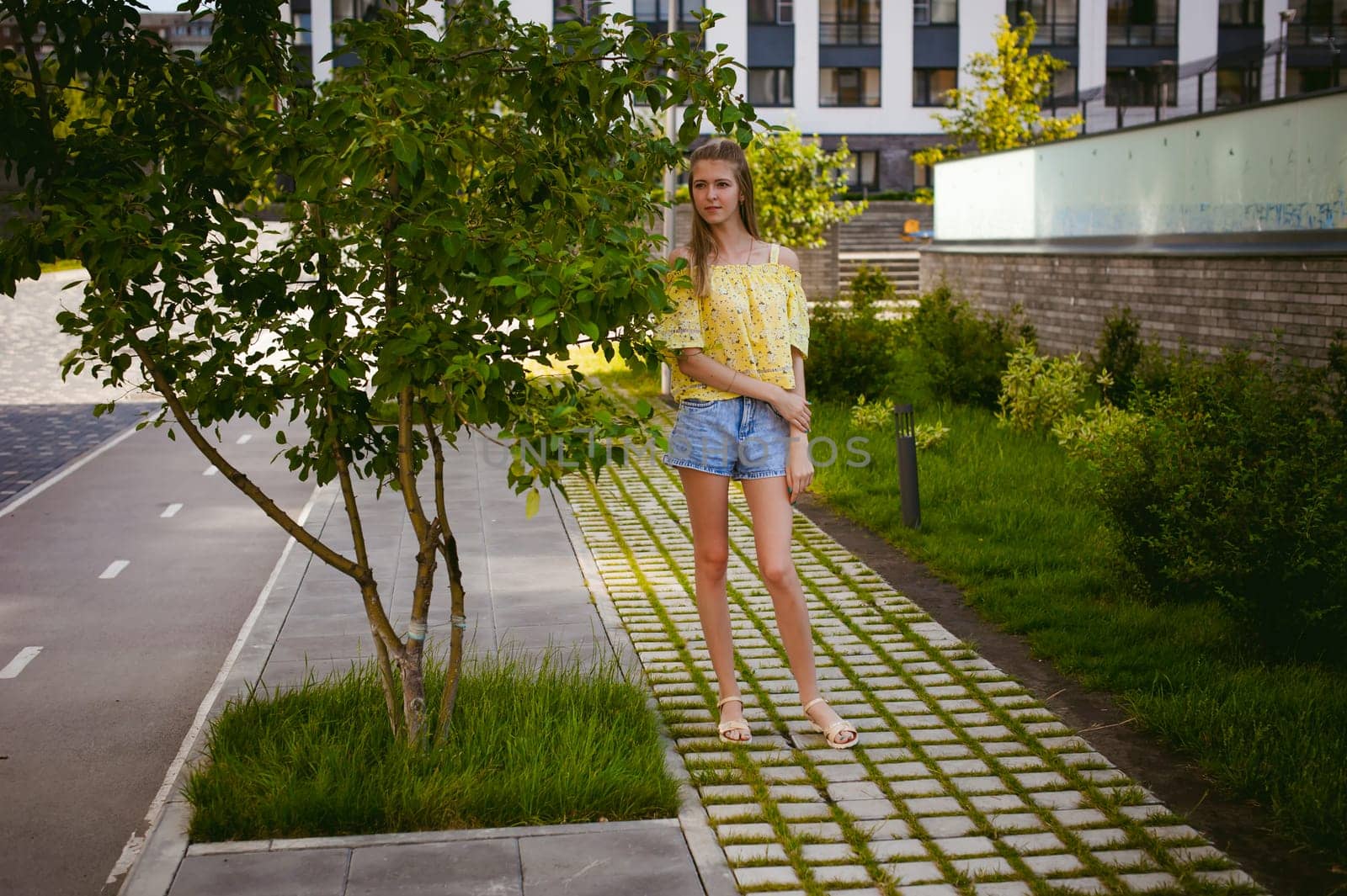Young beautiful woman is strolling outdoors in the yard, against the backdrop of greenery and houses, warm summer sunny day