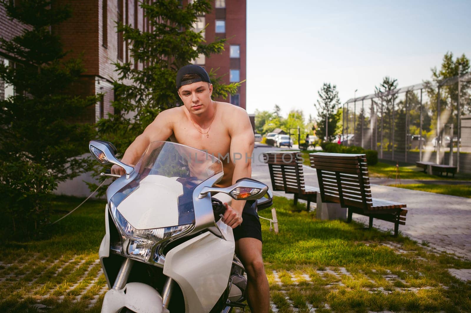 Handsome sexy male athlete man with naked torso in yard of house in parking lot sitting on a white sport motorcycle, an early sunny summer morning
