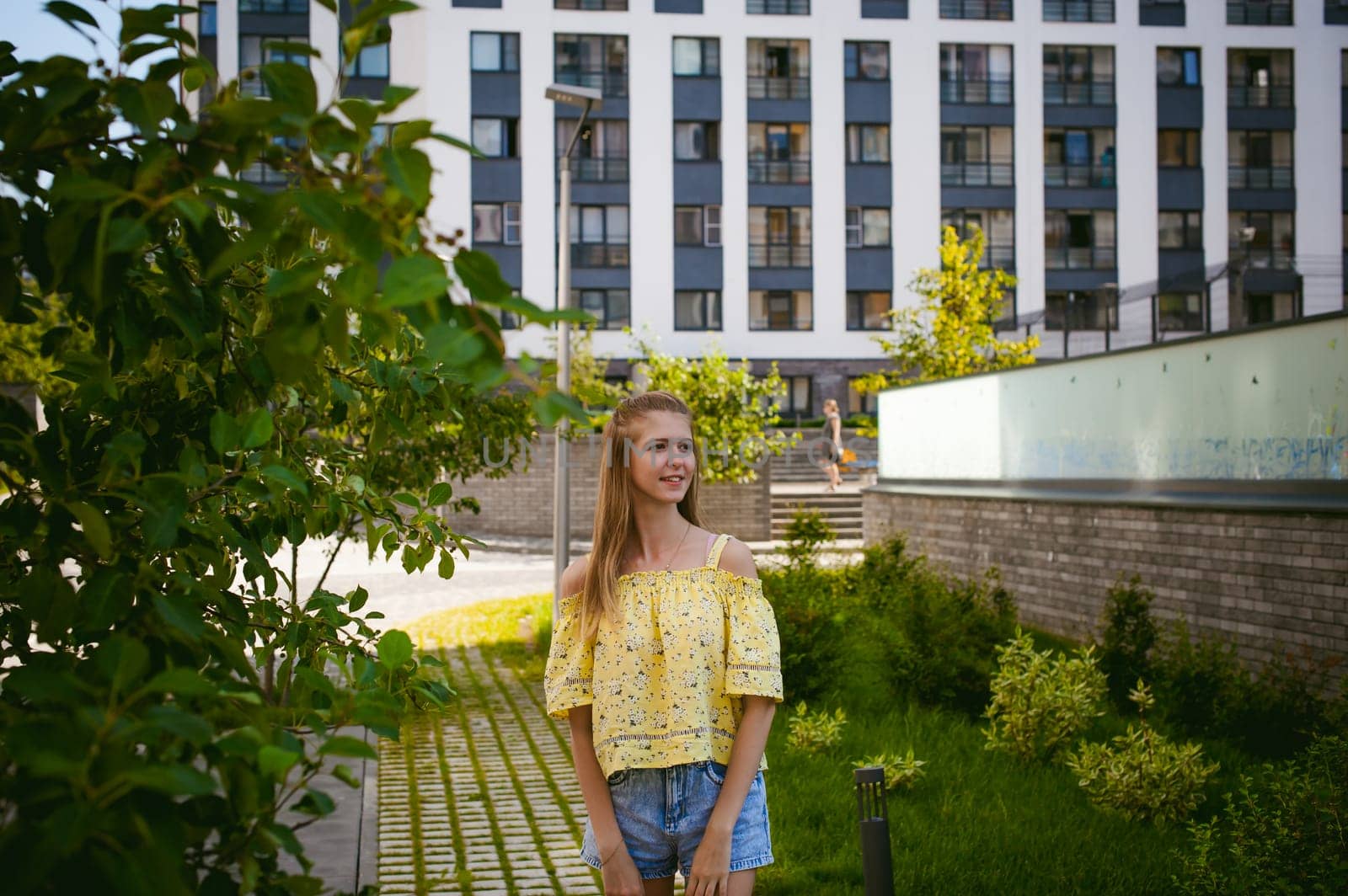 Young beautiful woman is strolling outdoors in the yard, against the backdrop of greenery and houses, warm summer sunny day