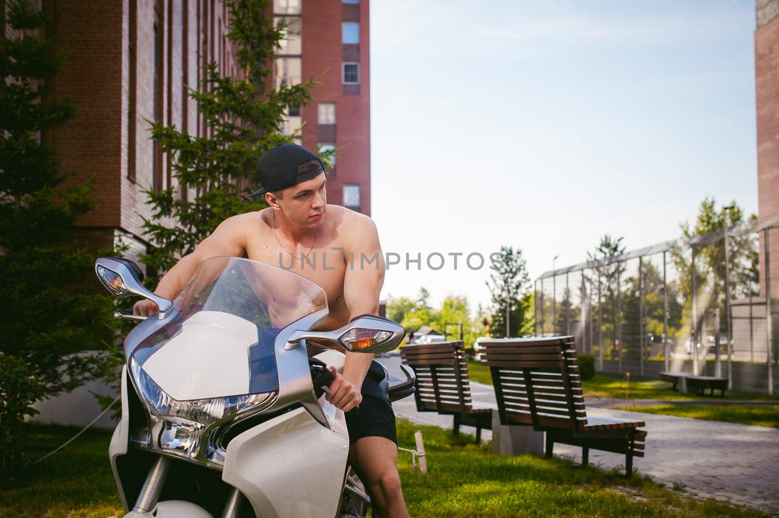 Handsome sexy male athlete man with naked torso in yard of house in parking lot sitting on a white sport motorcycle, an early sunny summer morning