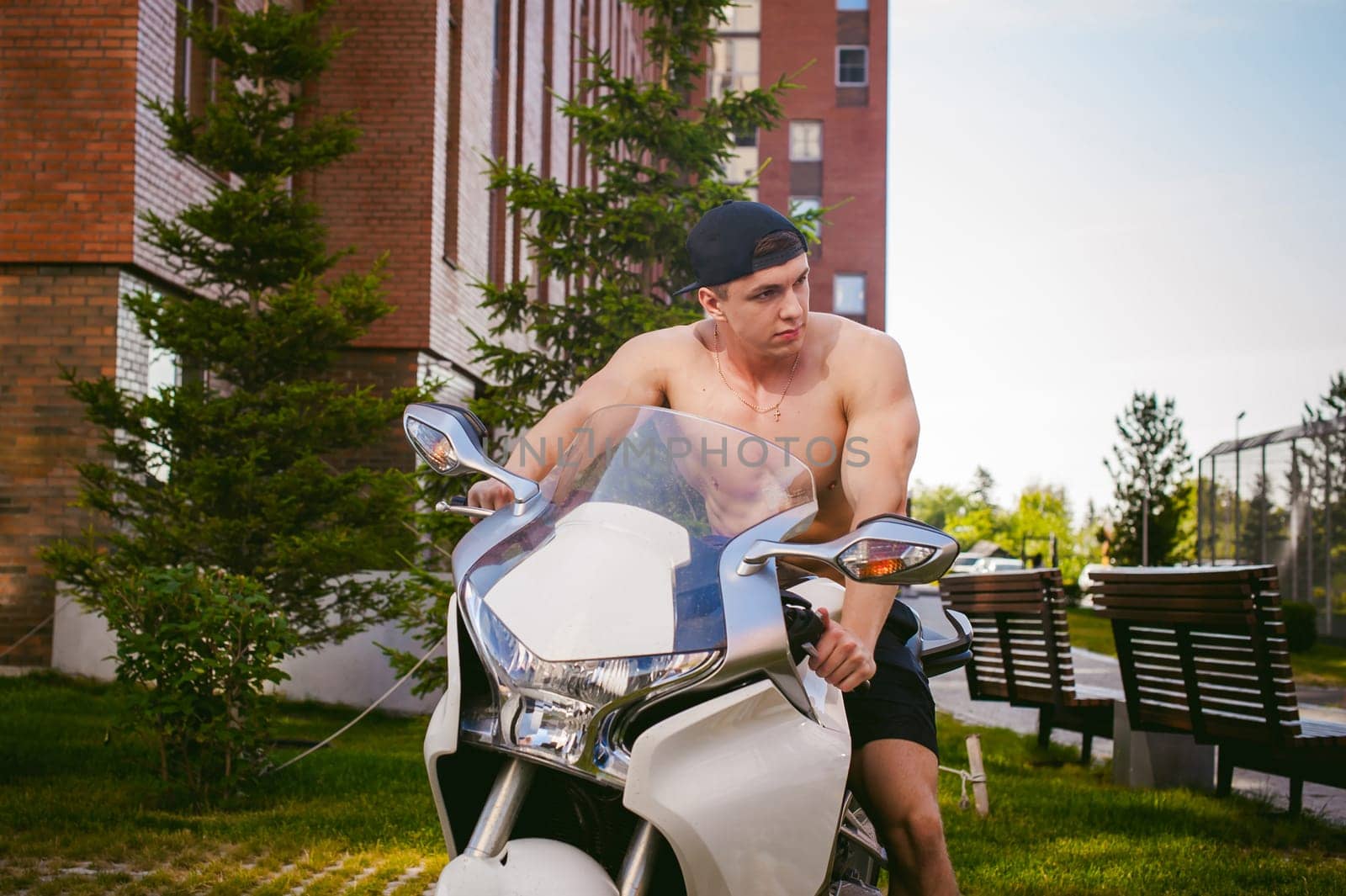 Handsome sexy male athlete man with naked torso in yard of house in parking lot sitting on a white sport motorcycle, an early sunny summer morning