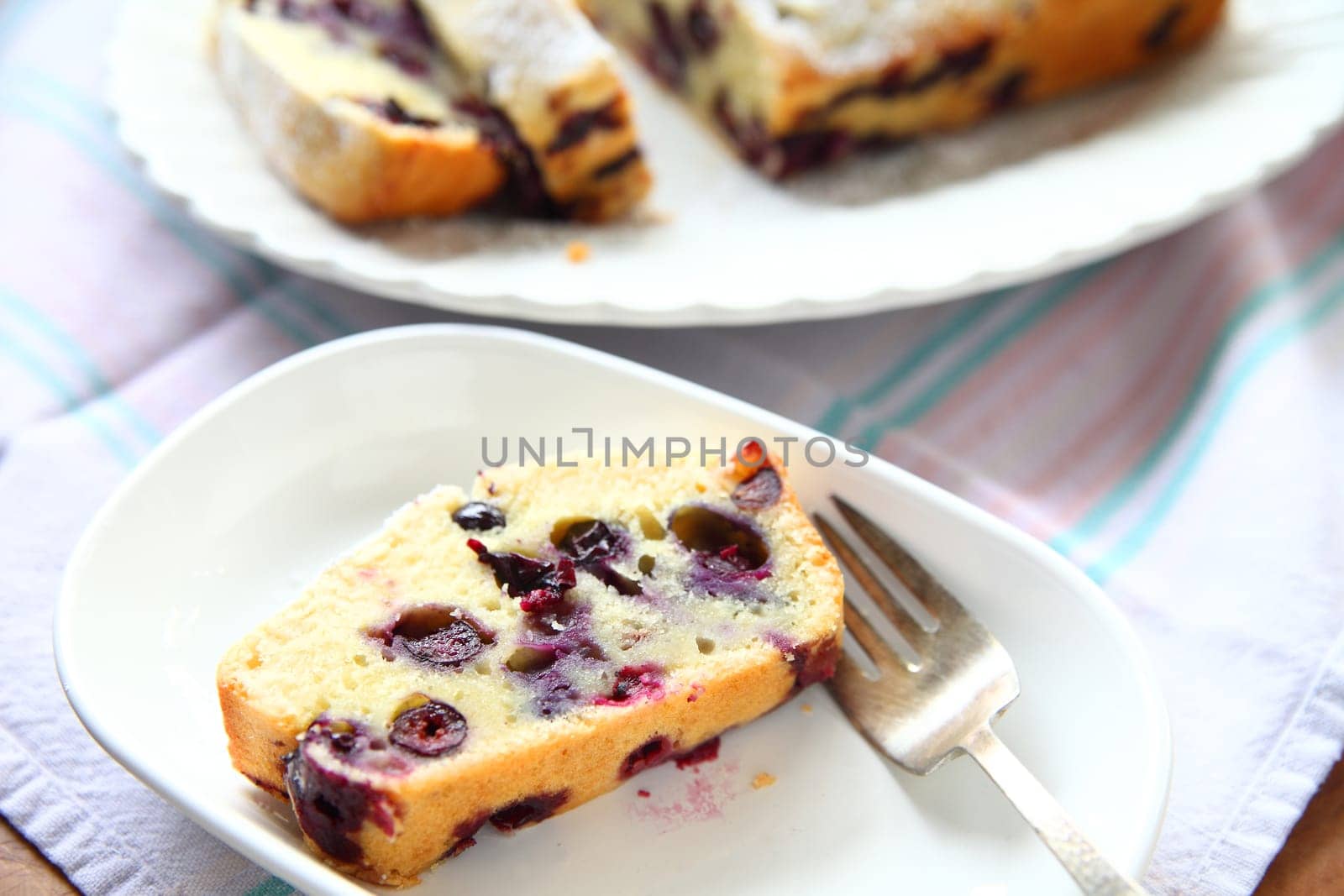 A serving of fresh blueberry cake on a white dish, rest of cake in background