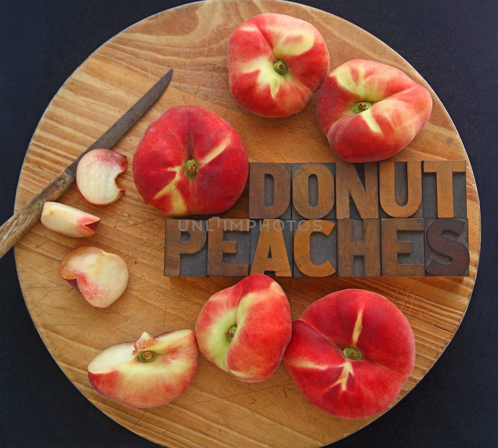 Fresh donut peaches, whole and with pieces cut, with words in wood type and a kitchen knife on a cutting board