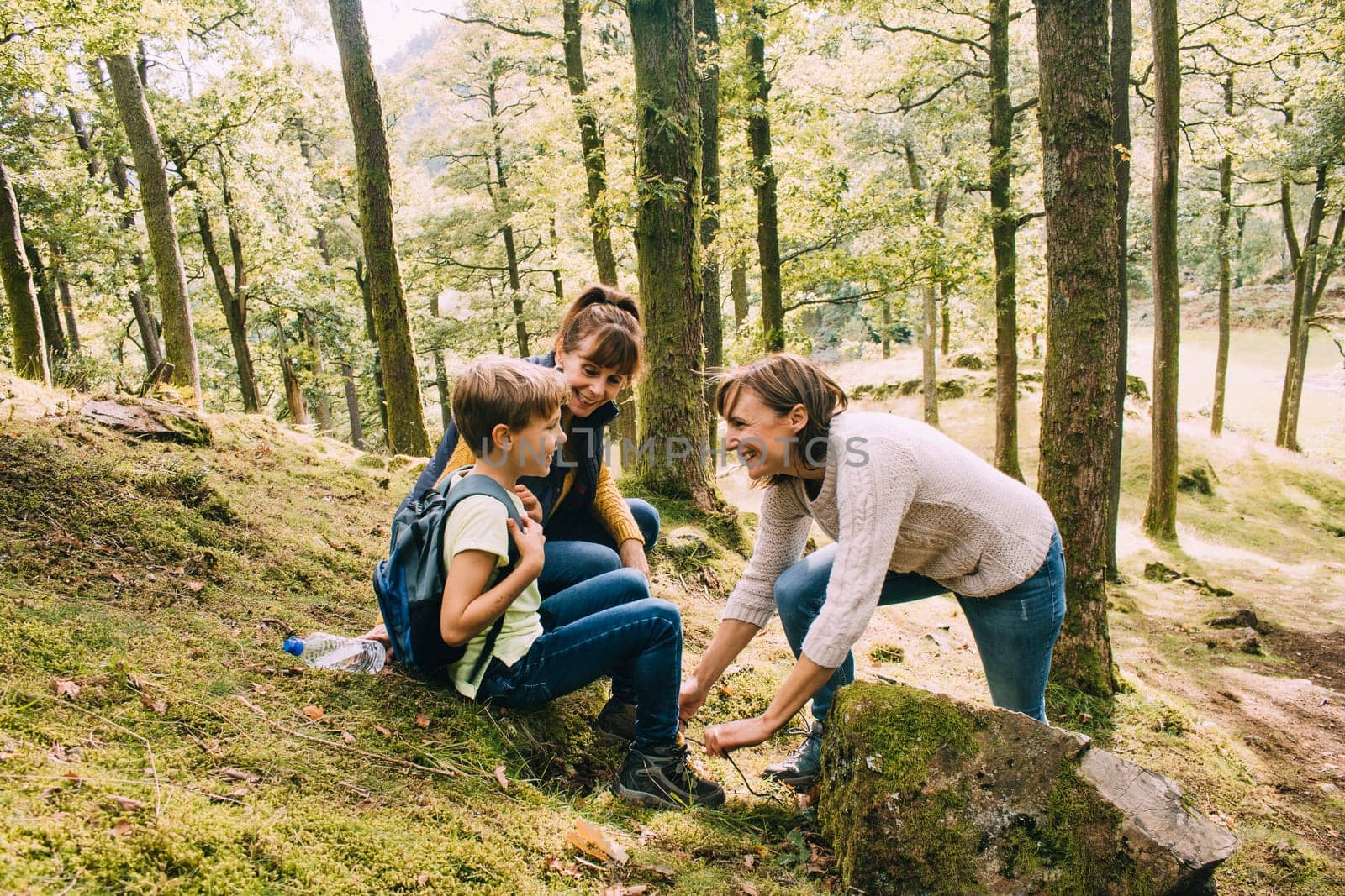 Happy family are hiking through the woodlands together. They have stopped to help the boy tie the laces on his hiking boots.