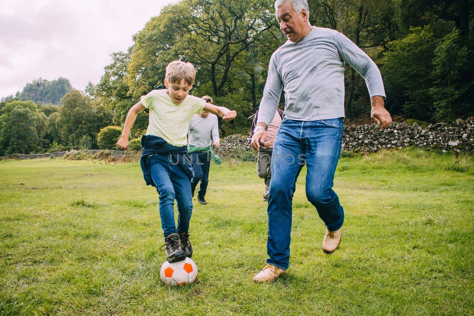 Three generation family are playing football together in a field. There are two boys, their father and their grandfather.