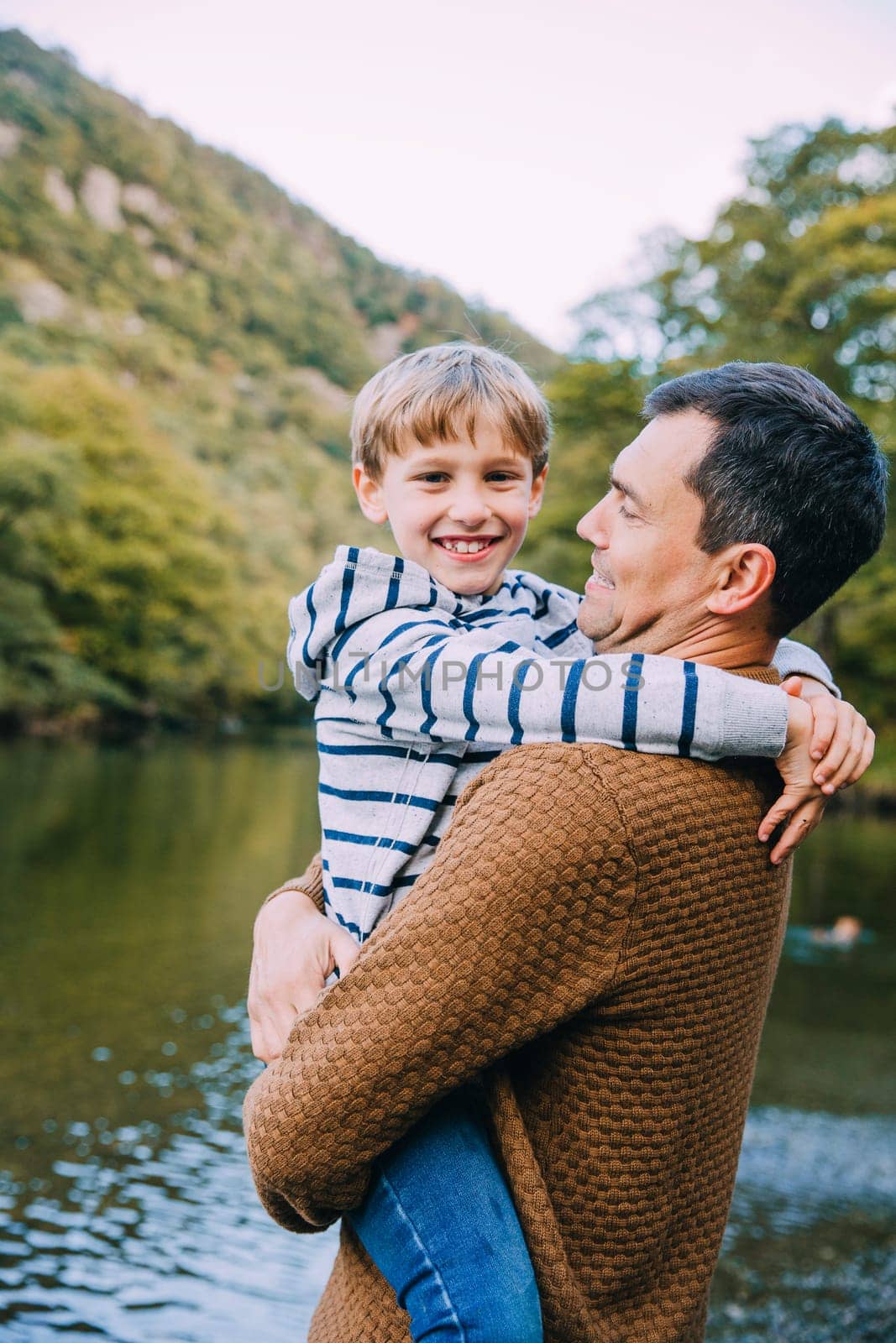 Little boy is smiling for the camera while being held by his father. They are standing near a lake.