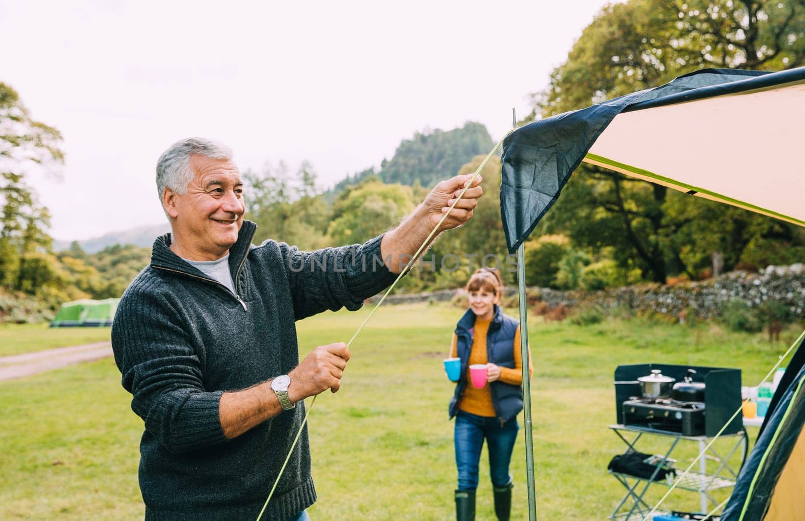 Senior couple have arrive at the campsite they are staing in. The man is putting up their tent while his wife makes cups of tea for everybody.