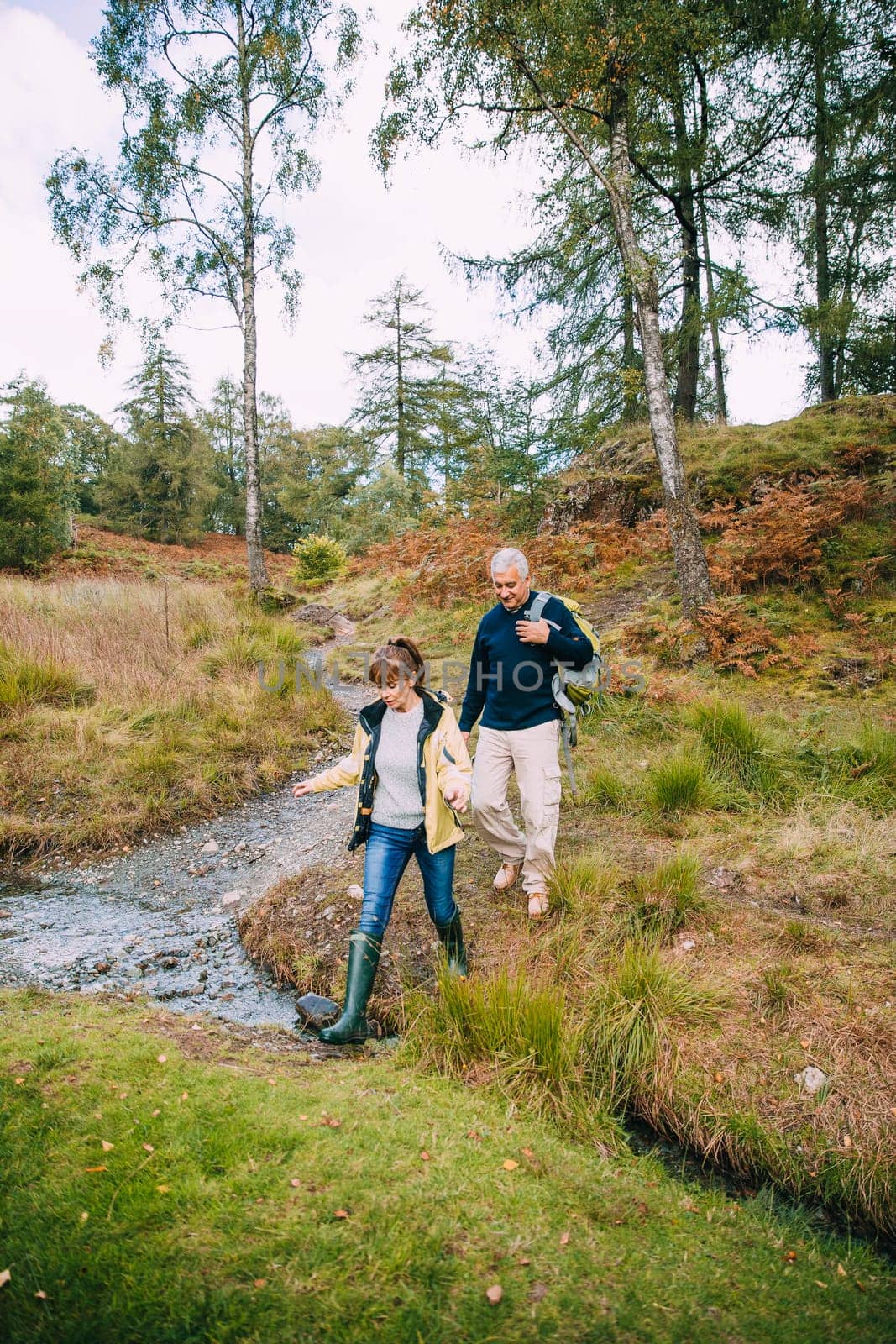 Senior couple are enjoying a walk through the woodlands together. They have come across a small stream which they are stepping over.