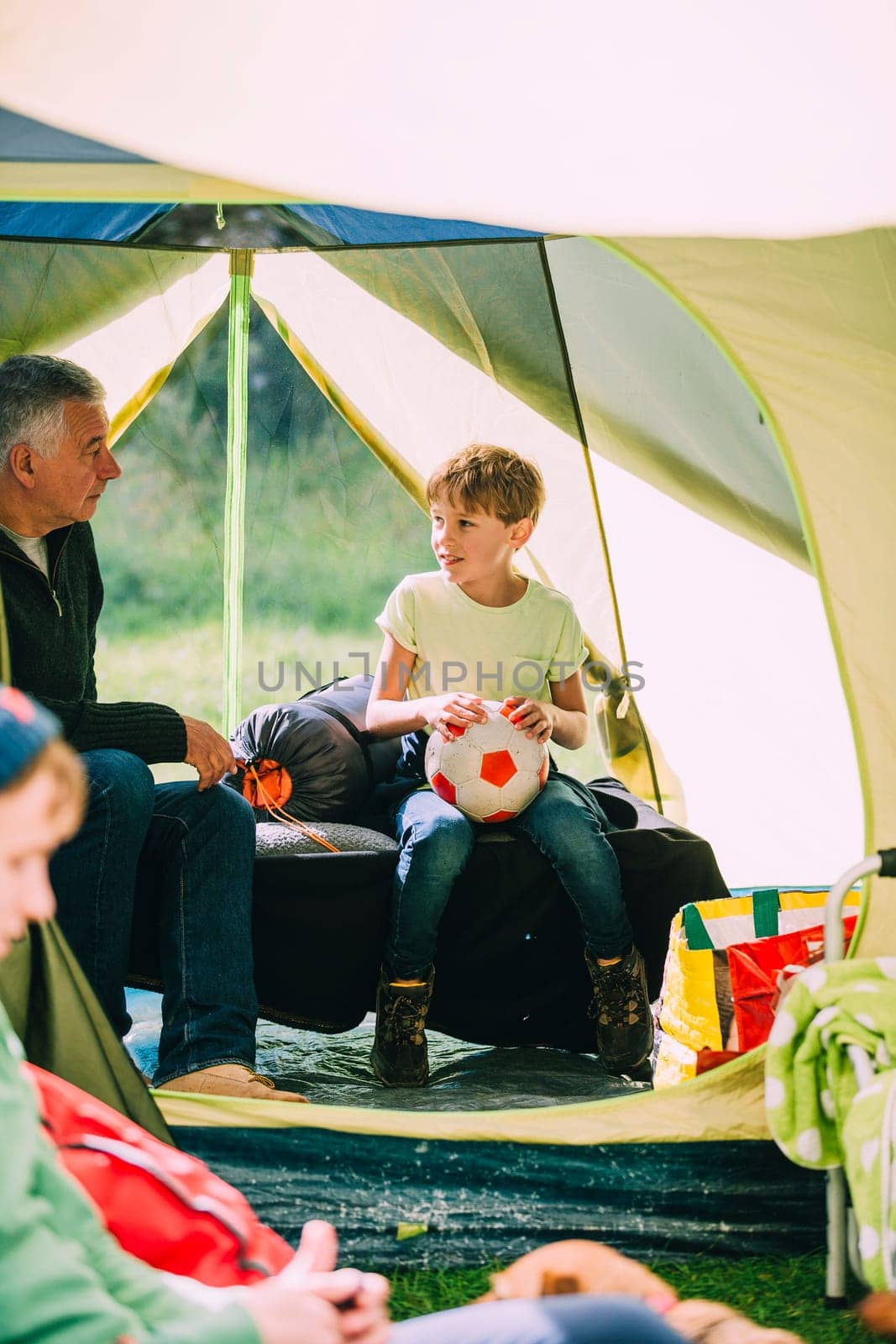 Little boy is sitting in a tent with his grandad after a game of football. He is holding the ball and they are talking.