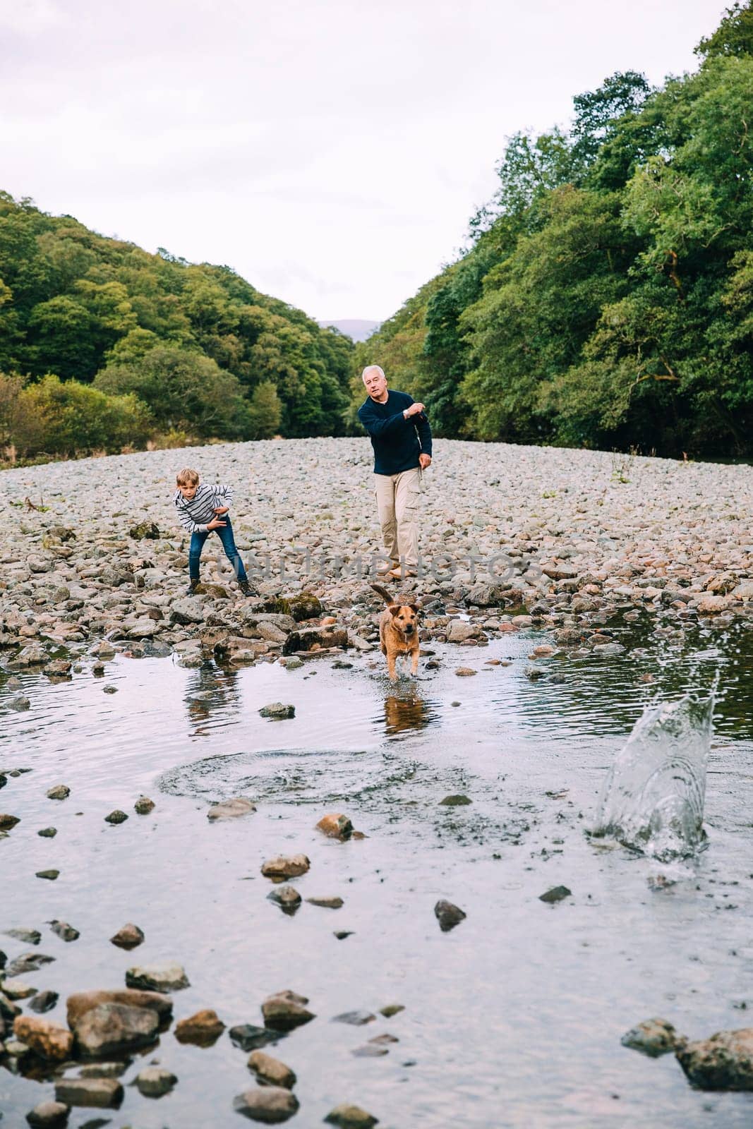 Little boy and his grandfather are out on a wlak in the woodlands. They have found a small pond and are throwing stones in for their pet dog to fetch.