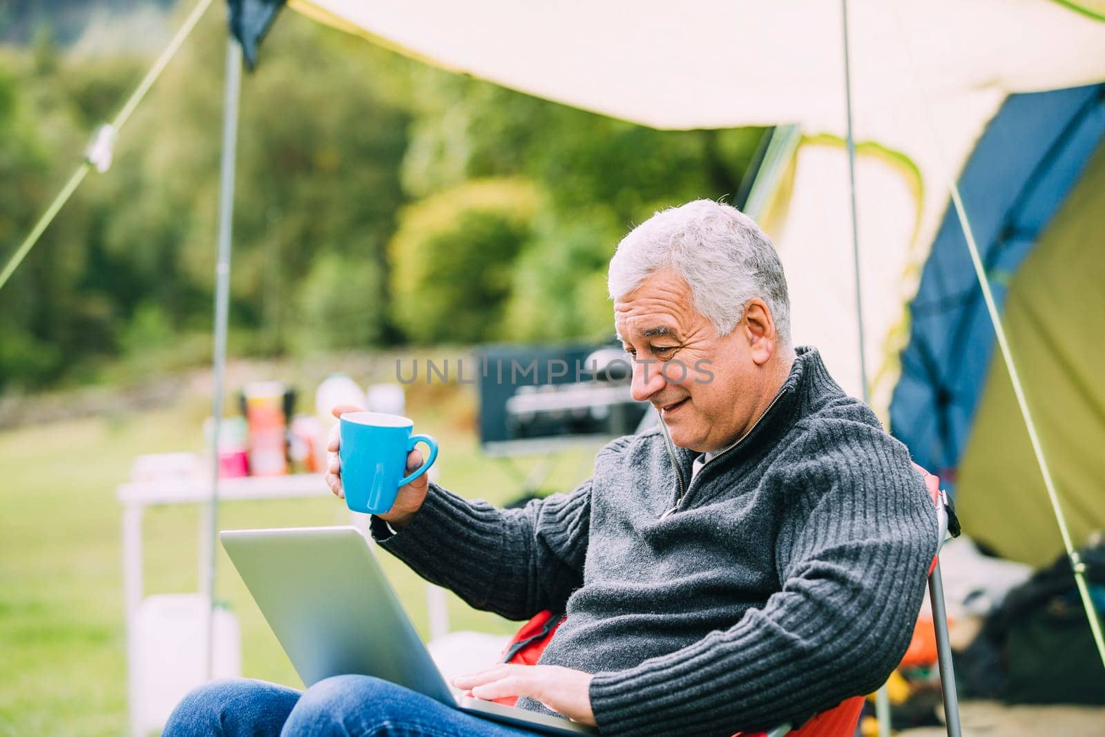 Senior man is relaxing in a chair by his tent in the campsite. He is enjoying a cup of tea while using a laptop.