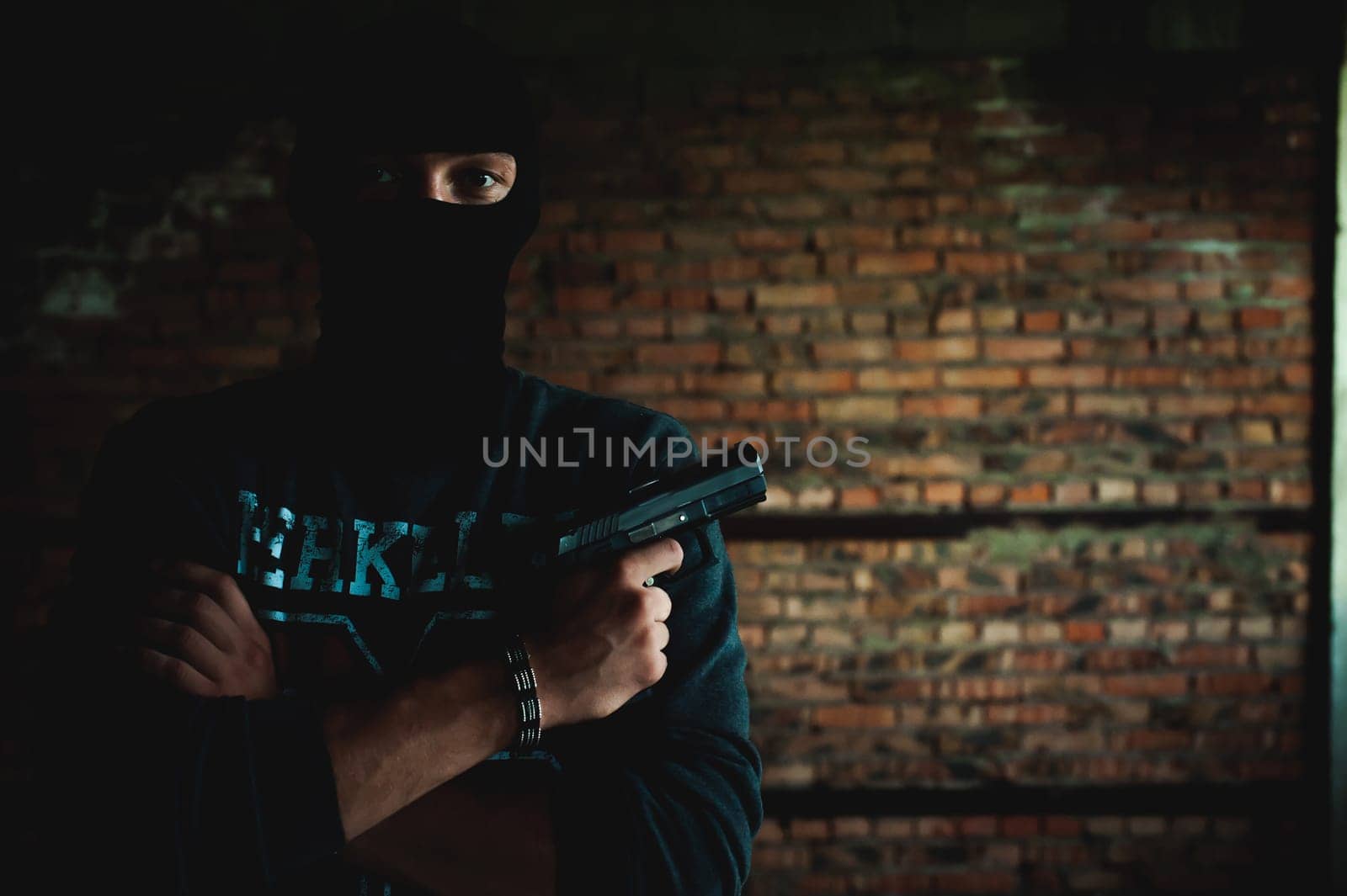 Athletic man in a balaclava, ski mask on his head, holds a gun in his hand, with his arms crossed over his chest