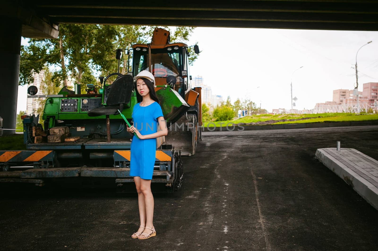 Asian woman in a blue dress and white protective helmet. engineer supervises inspection and control of works at construction site. on background of road construction machinery