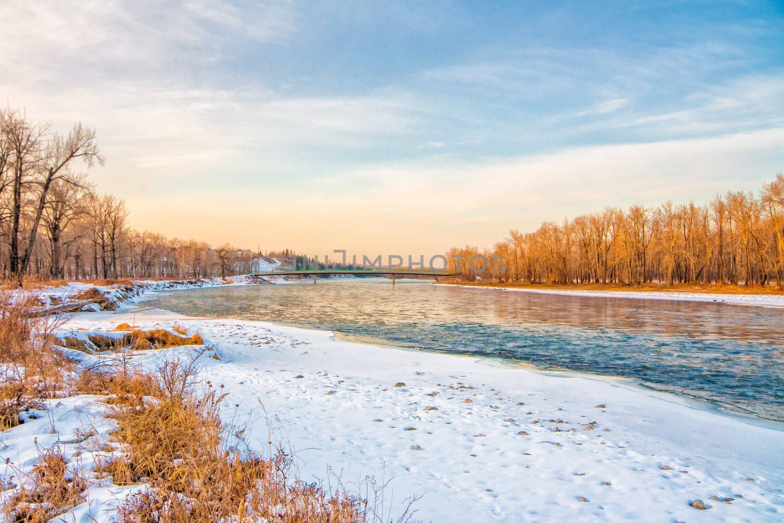 Landscape of the Bow River with snow covered banks, warm light and a bridge in the distance.