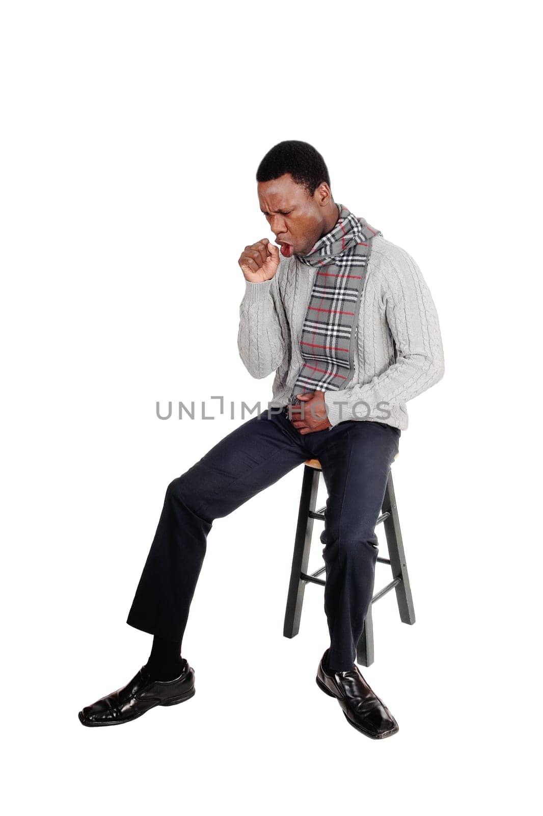 A sick African American man in a gray sweater and scarf sitting on a.chair and coughing, isolated for white background.