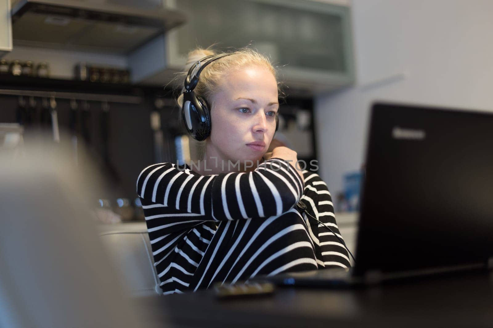 Woman in her casual home clothing working and studying remotely from her small flat late at night. Home kitchen in background. Great flexibility of web-based courses and study programmes.