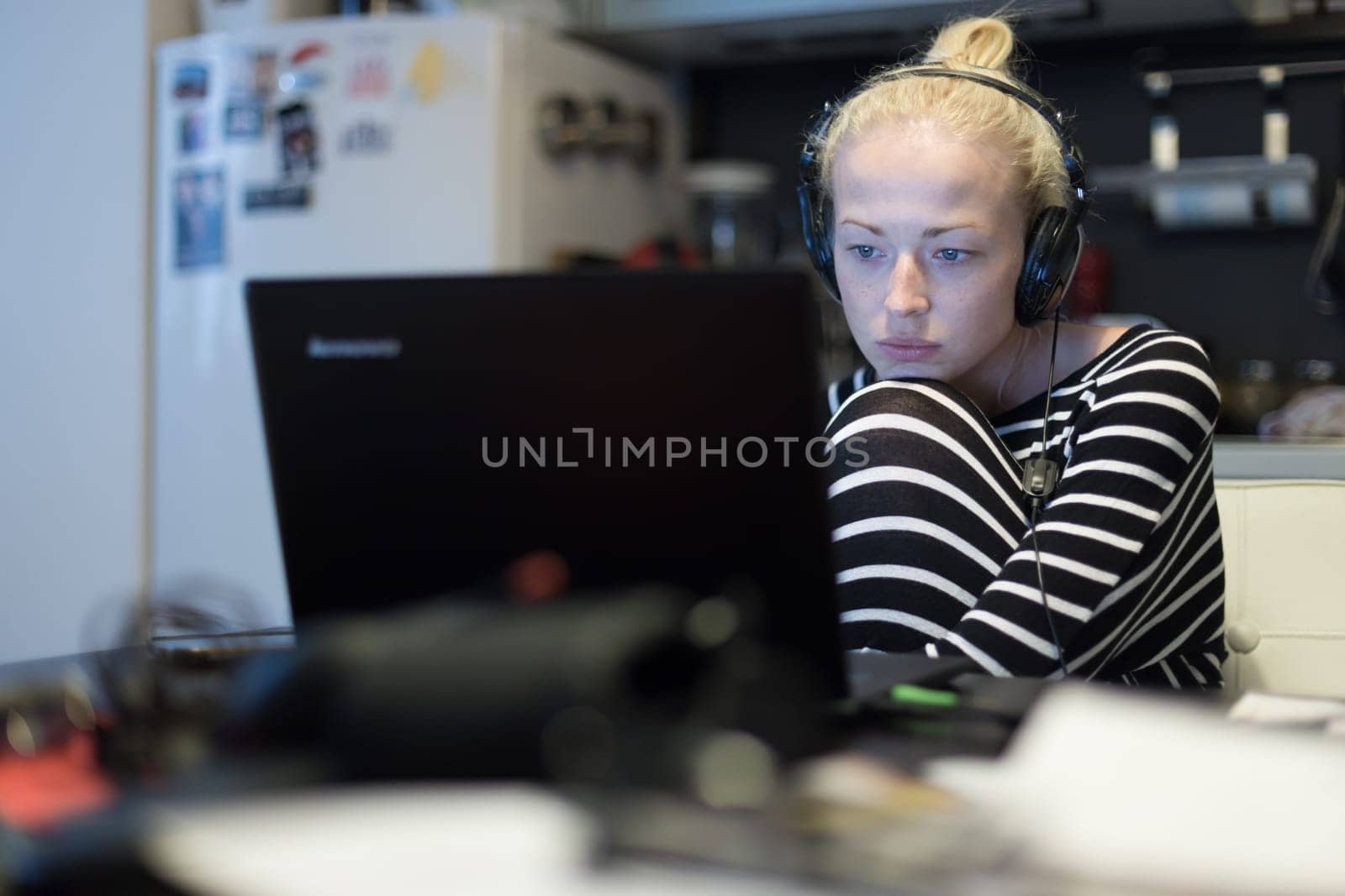 Woman in her casual home clothing working and studying remotely from her small flat late at night. Home kitchen in background. Great flexibility of web-based courses and study programmes.