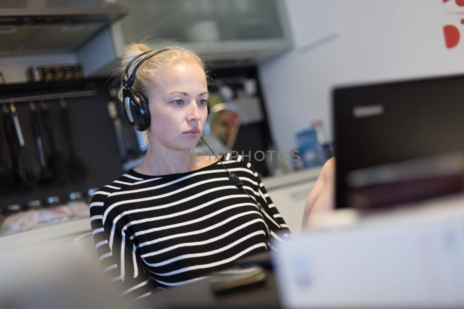 Woman in her casual home clothing working and studying remotely from her small flat late at night. Home kitchen in background. Great flexibility of web-based courses and study programmes.