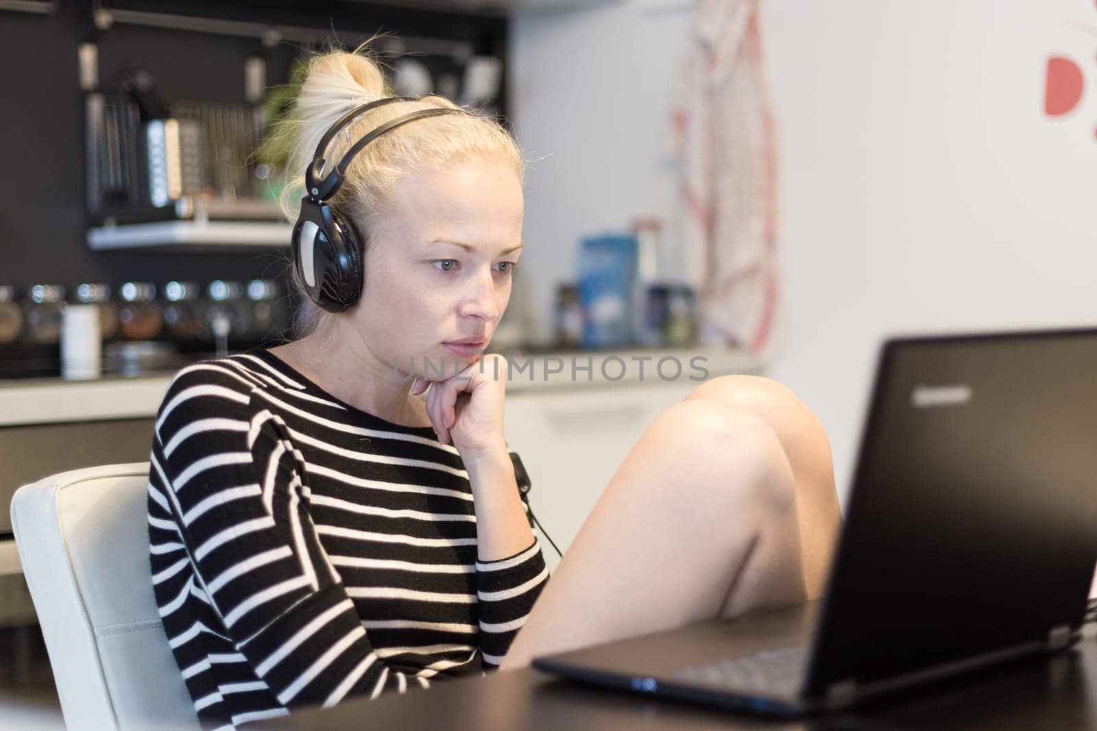Woman in her casual home clothing working and studying remotely from her small flat late at night. Home kitchen in background. Great flexibility of web-based courses and study programmes.