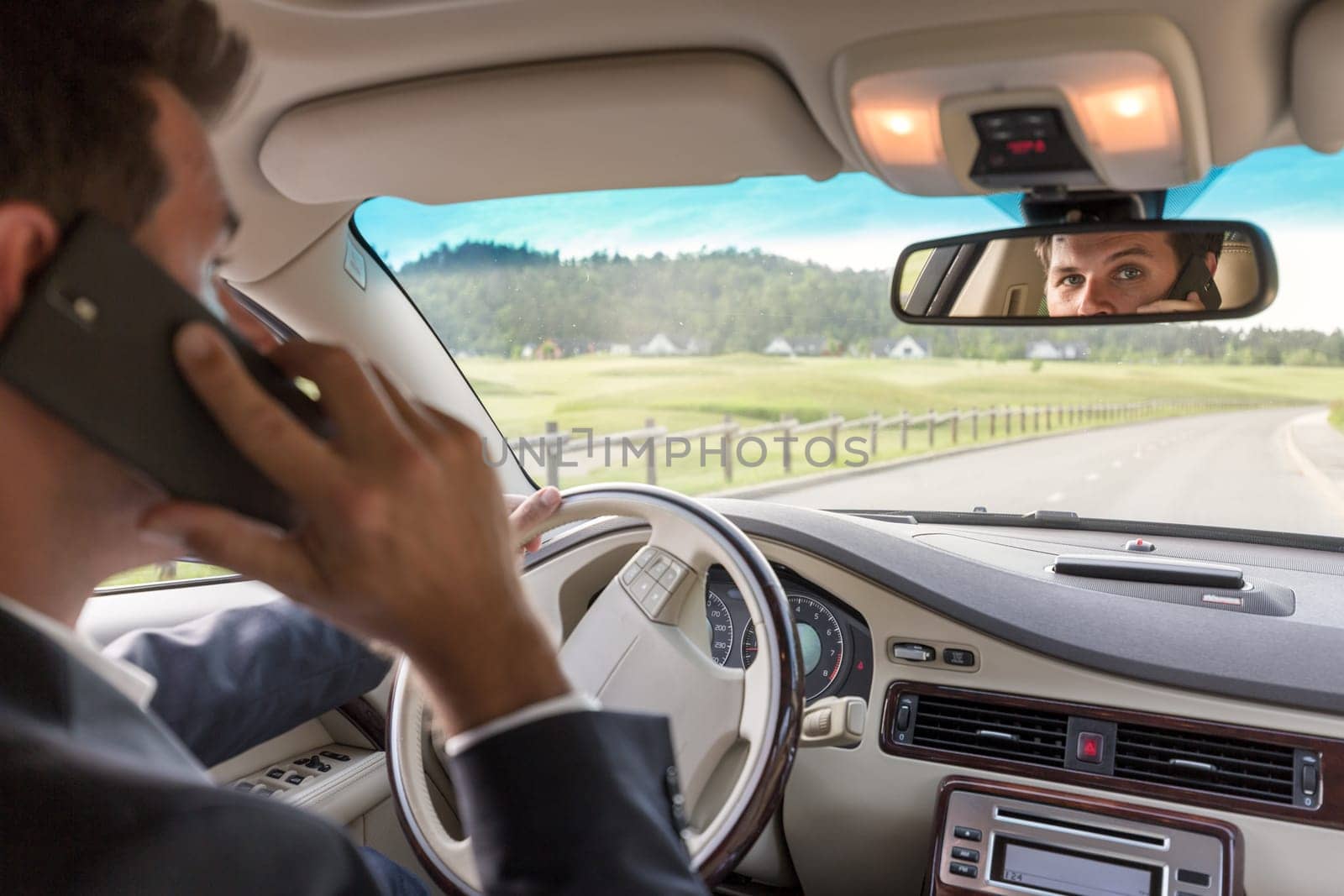 Businessman talking on cell phone while driving not paying attention to the road. Focus on the man face in rearview mirror.
