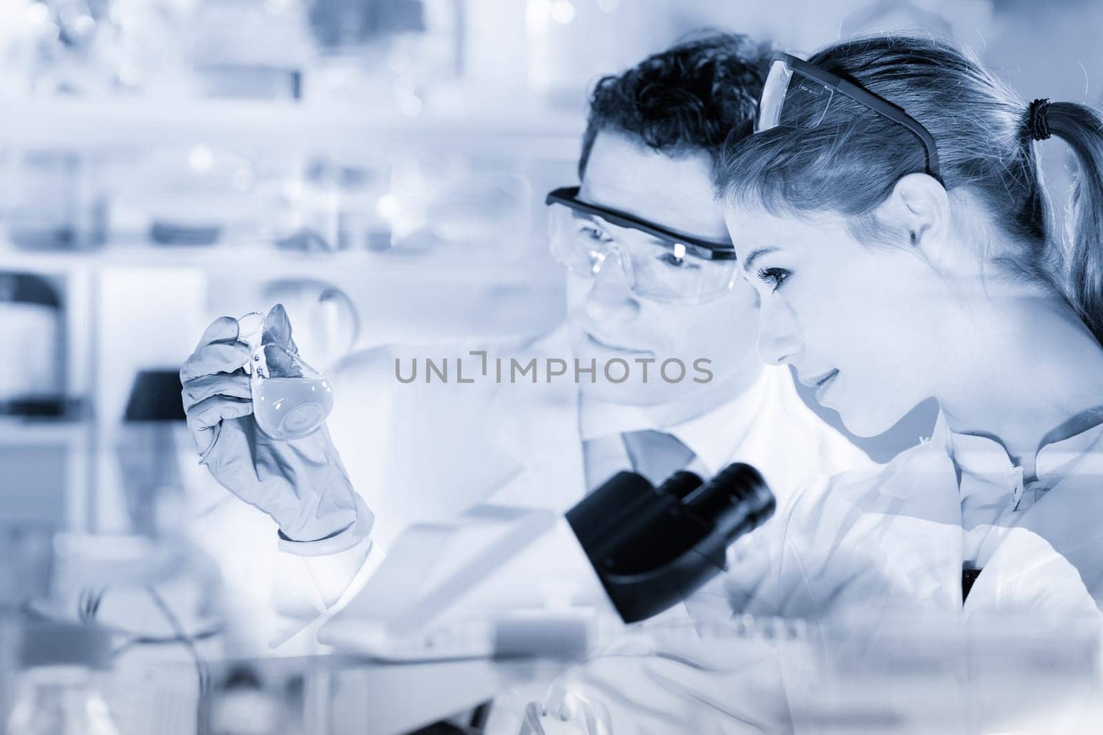 Chemical laboratory. Attractive young PhD student and her post doctoral supervisor scientist observing the blue indicator color shift after the solution destillation. Greyscale blue toned image.