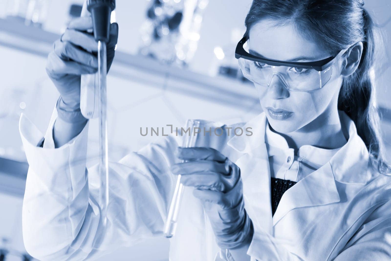 Life scientists researching in laboratory. Focused female life science professional pipetting solution into glass cuvette. Healthcare and biotechnology. Greyscale blue toned image.