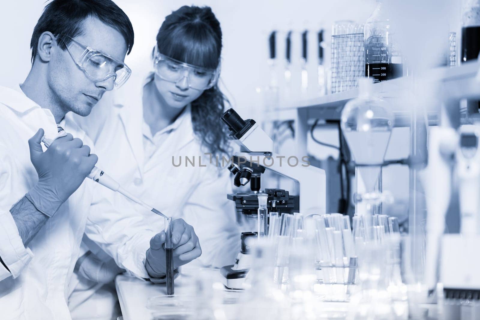 Young male researcher pipetting red liquid in the glass tube in the laboratory. Female asistant science student watching and learning the protocol.