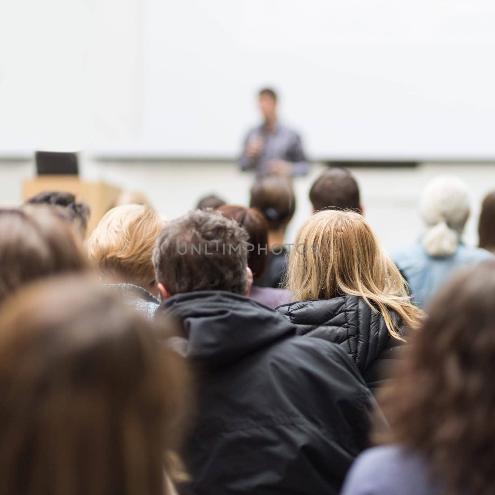 Male speaker giving presentation in lecture hall at university workshop. Audience in conference hall. Rear view of unrecognized participant in audience. Scientific conference event.