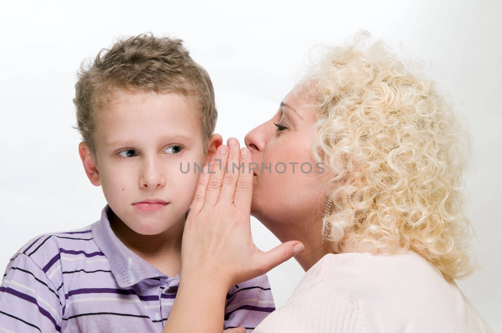 Side head-and-shoulder view of a blond curly woman into the ear of an 8-year-old boy whispering at gunpoint in front of a white background.