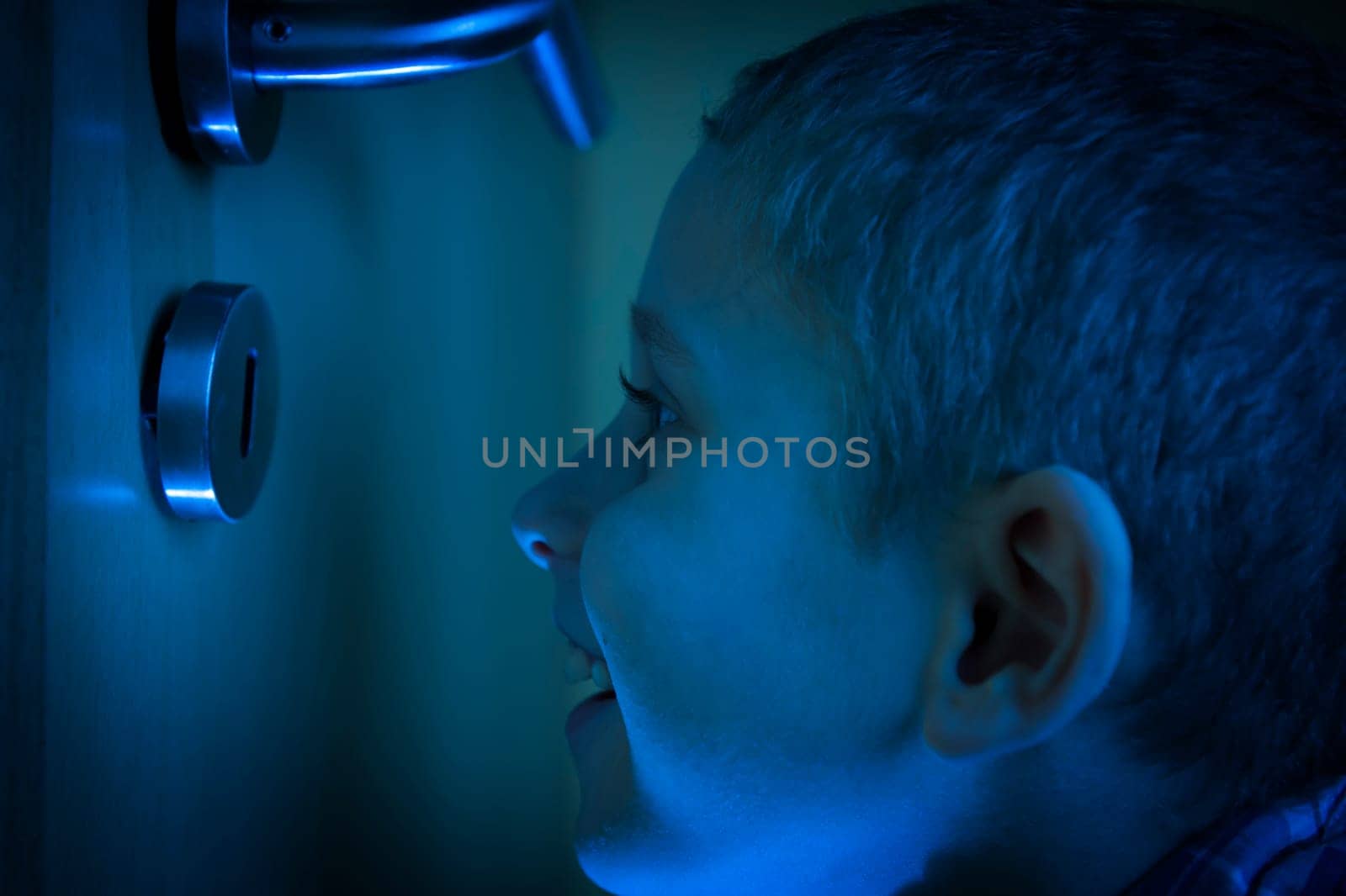 Boy in the dark room looks through a keyhole in the glow of the flashlight.