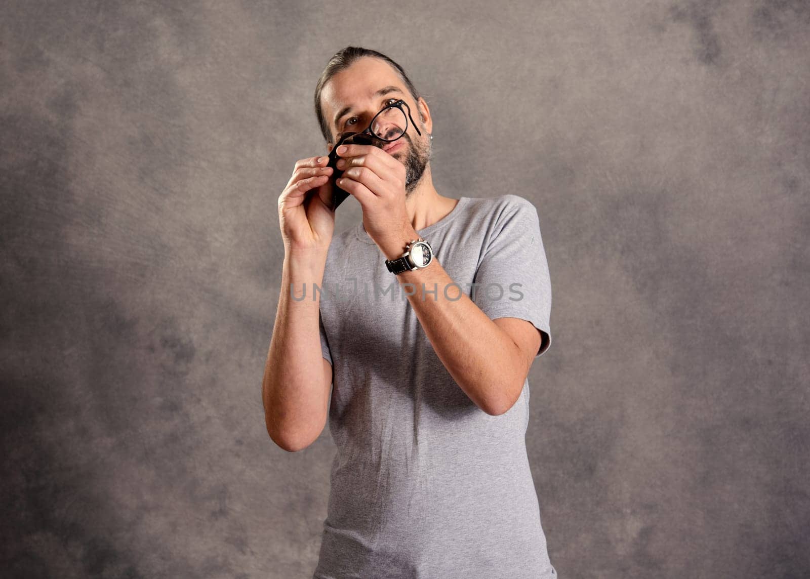 young bearded man ceaning his glasses in front of gray background