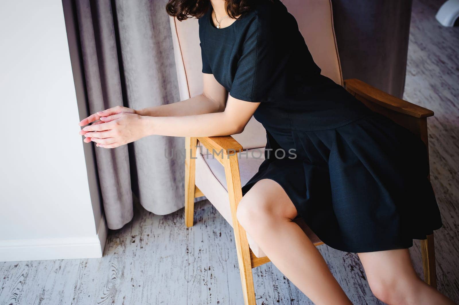 Young attractive woman with long legs in black elegant dress, sits in chair near window in interior of room.