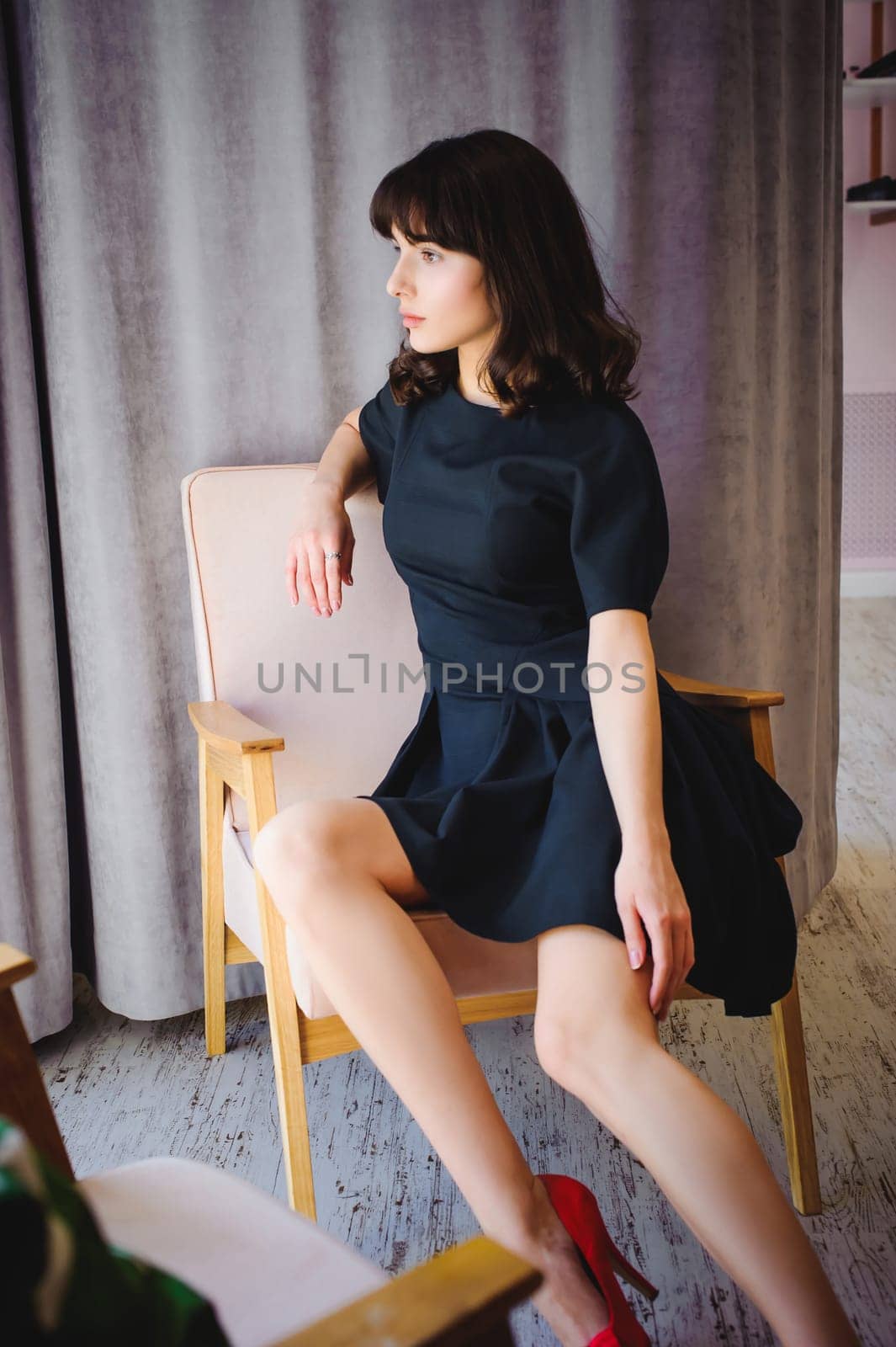 Young attractive woman with long legs in black elegant dress, sits in chair near window in interior of room.