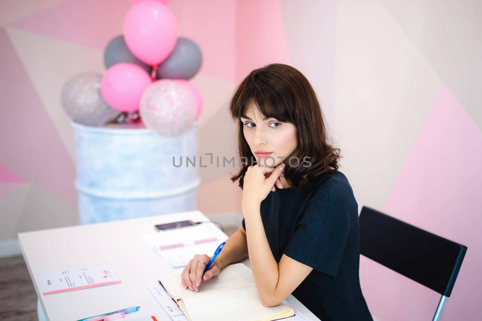 Portrait of a beautiful business woman making notes in a notebook. Holds a pen, sitting in the workplace in a pink office