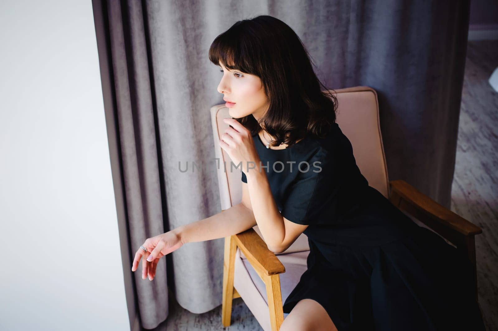 Young attractive woman with long legs in black elegant dress, sits in chair near window in interior of room.
