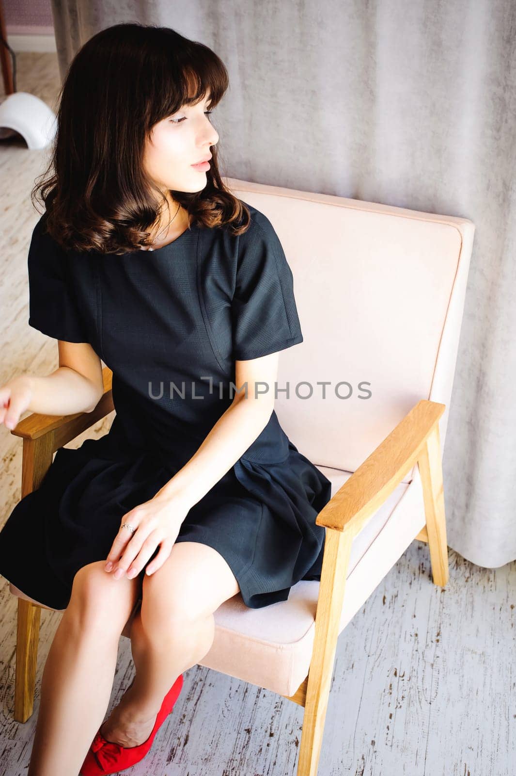 Young attractive woman with long legs in black elegant dress, sits in chair near window in interior of room. Seductive shoes of red light, on beautiful legs