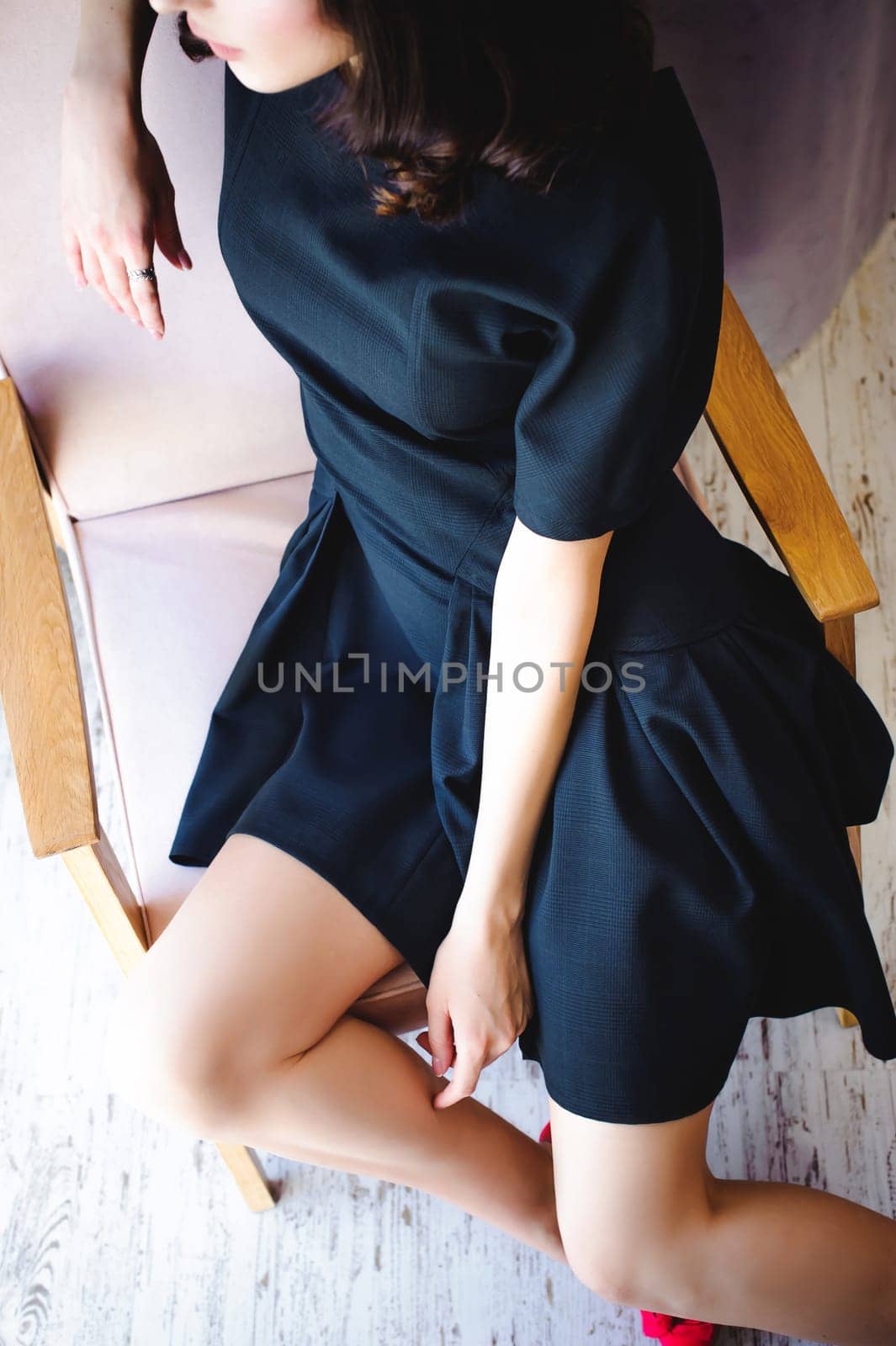 Young attractive woman with long legs in black elegant dress, sits in chair near window in interior of room.